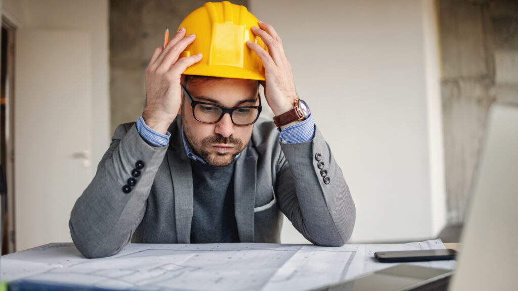 Focused architect holding his head, sights and looking at blueprints. Something got wrong.
